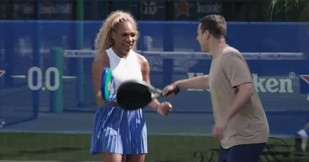 Serena Williams lacht op een padelbaan met een padelracket, Heineken 0.0 branding op de achtergrond